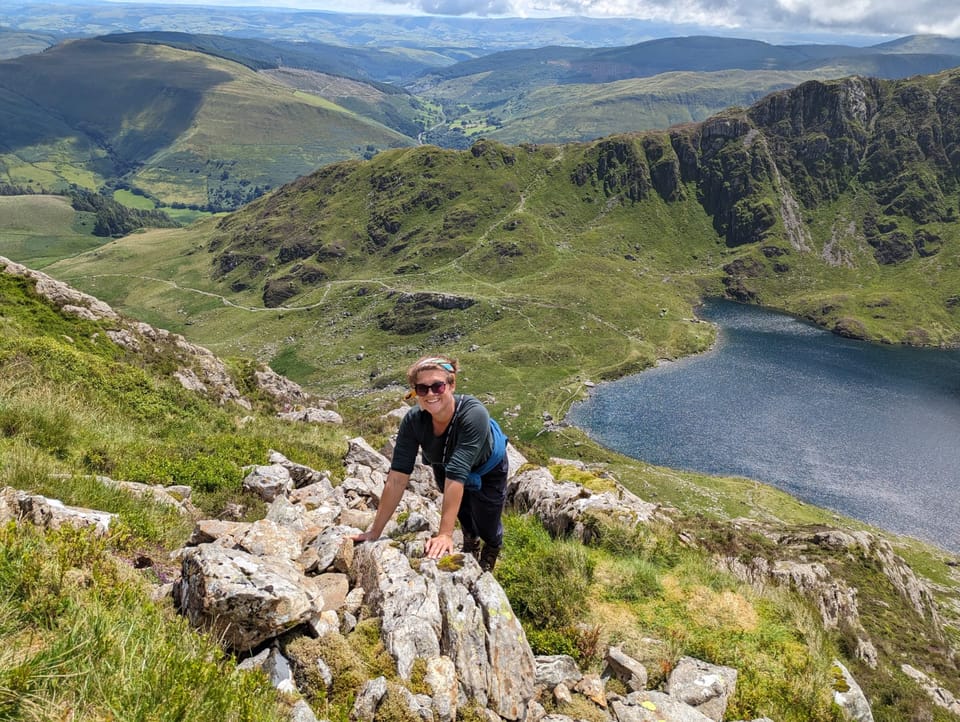 A smiling woman on the side of a mountain, with views of mountains and a lake